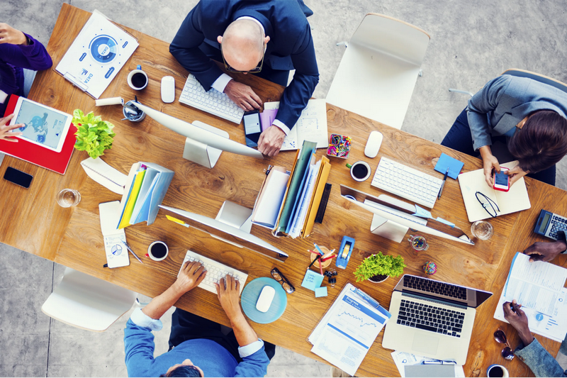 An overhead view of a group of people working together at a cluttered office desk with computers and various work materials.