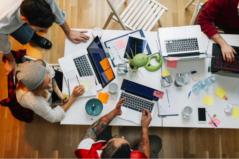 A group of people working together at a cluttered table with laptops and office supplies.