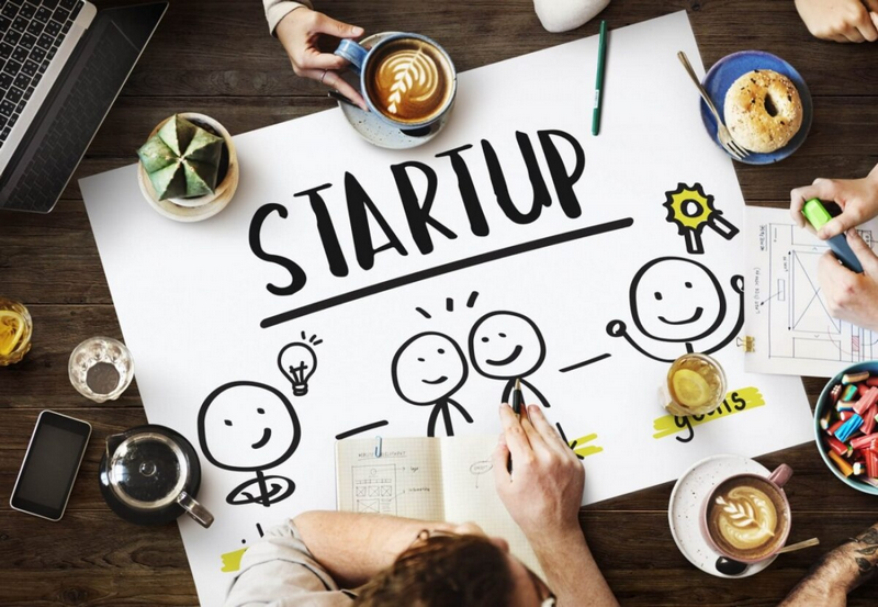 A group of people brainstorming around a table with a large paper labeled STARTUP and drawings of stick figures.